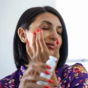 Woman applying cream on her face holding a serum bottle